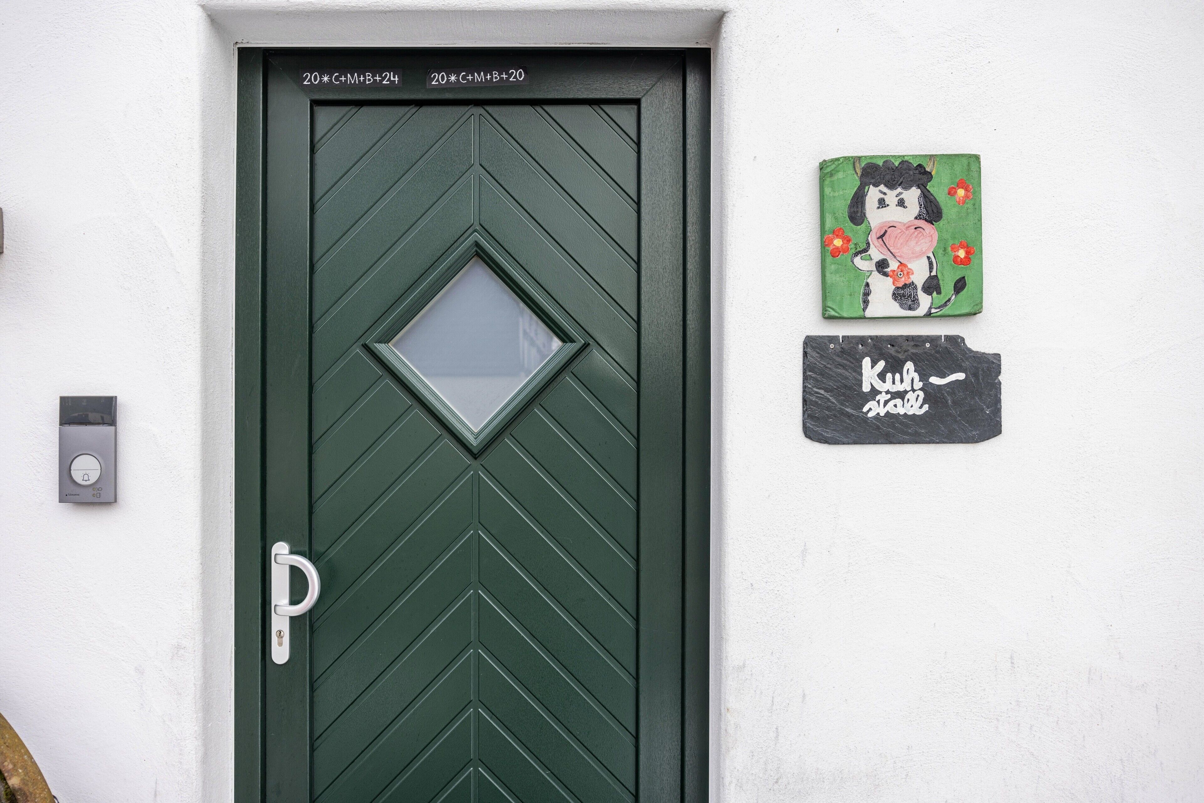 Une porte verte ornée d’une fenêtre en forme de losange, d’une sonnette et de deux décorations murales : l’une représente une vache et des fleurs, et l’autre porte l’inscription « Kuh Kuh ».