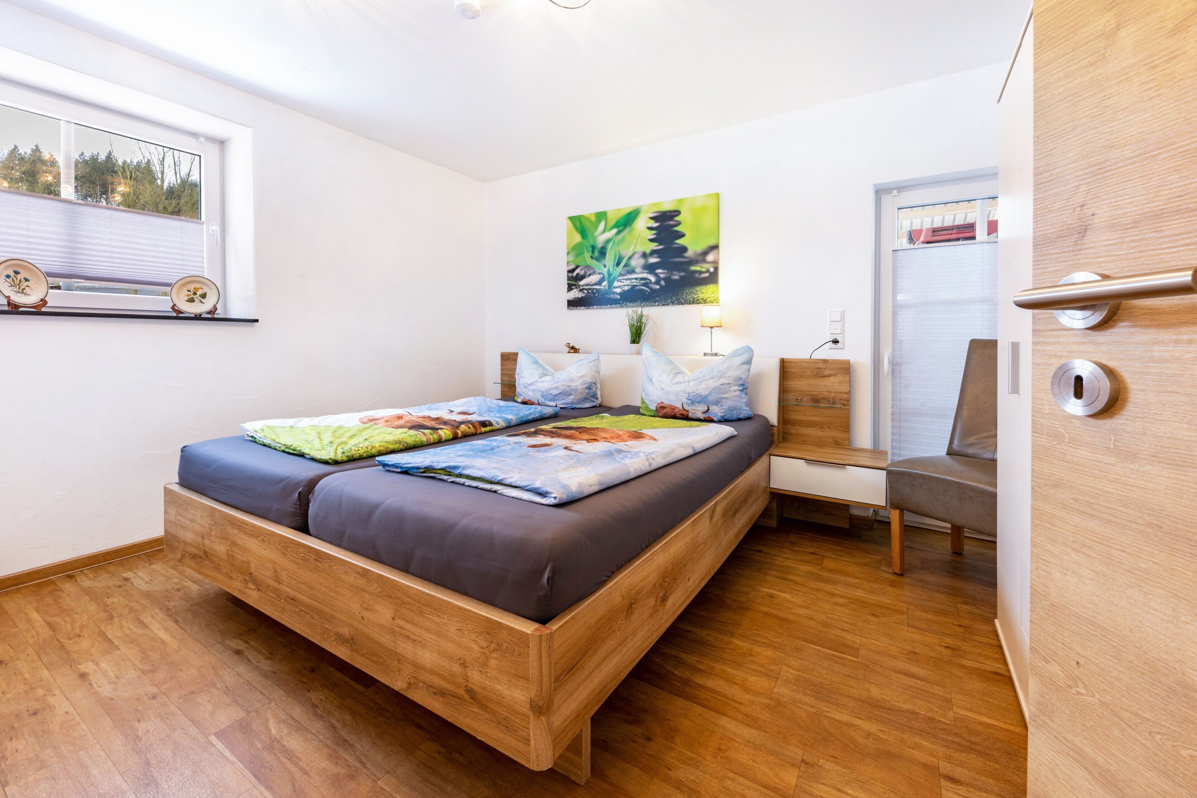 A bedroom with a wooden bed, a window with blinds, a painting on the wall, and a door with a handle.