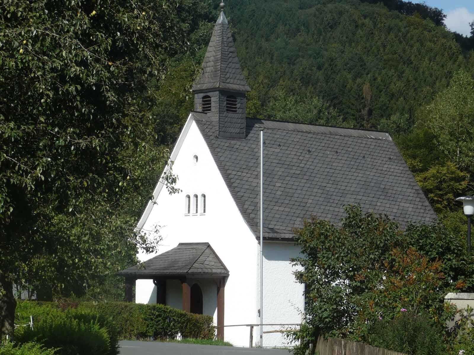 A white church with a steeple, surrounded by greenery and a mountain in the background.