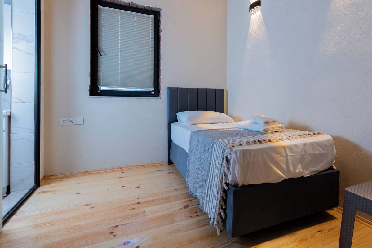 A small, neatly arranged bedroom with a bed, a bench, a window with blinds, and a wall-mounted light fixture.