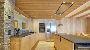 A modern kitchen with wooden cabinetry, a central island, a stone accent wall, and built-in appliances. - Chalet Almglück by we rent (Saalbach-Hinterglemm)