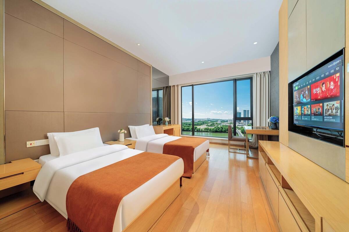 Habitación de hotel con dos camas, televisión, escritorio y ventana grande con vista a la ciudad.
