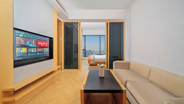 A modern living room with a flat-screen TV mounted on the wall, a sofa, a coffee table, and a view of a cityscape through large windows. - Xilaideng Guangzhou Service Apartment (Guangzhou)