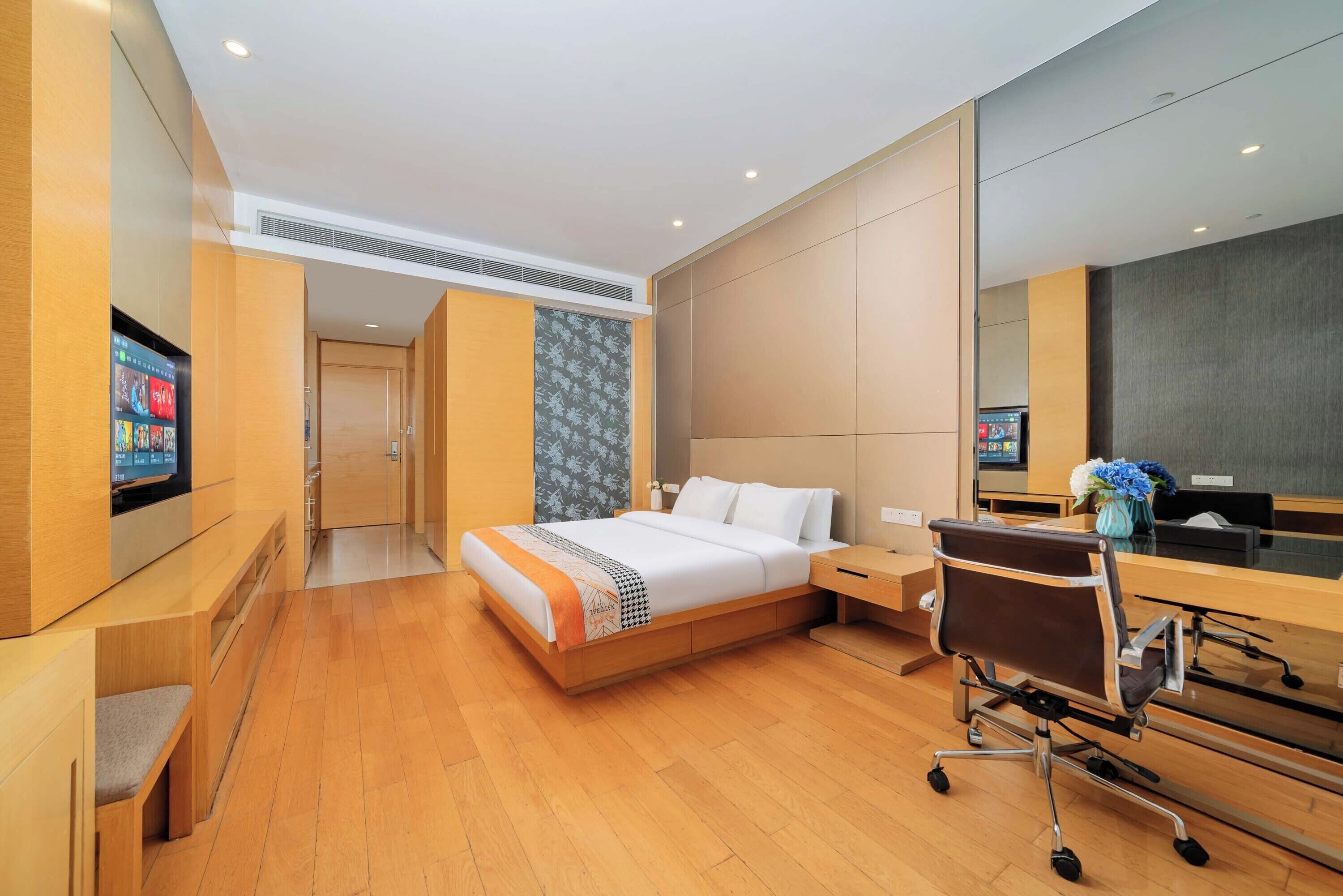 A modern hotel room with a wooden floor, a bed with a white and orange bedspread, a desk with a chair, and a large mirror.