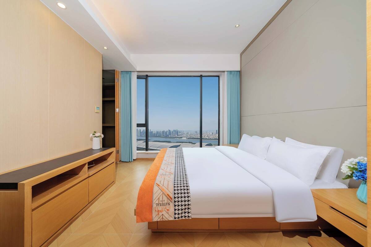 Una habitación de hotel moderna con una cama grande, una mesita de madera y vistas de la ciudad a través de un ventanal amplio.