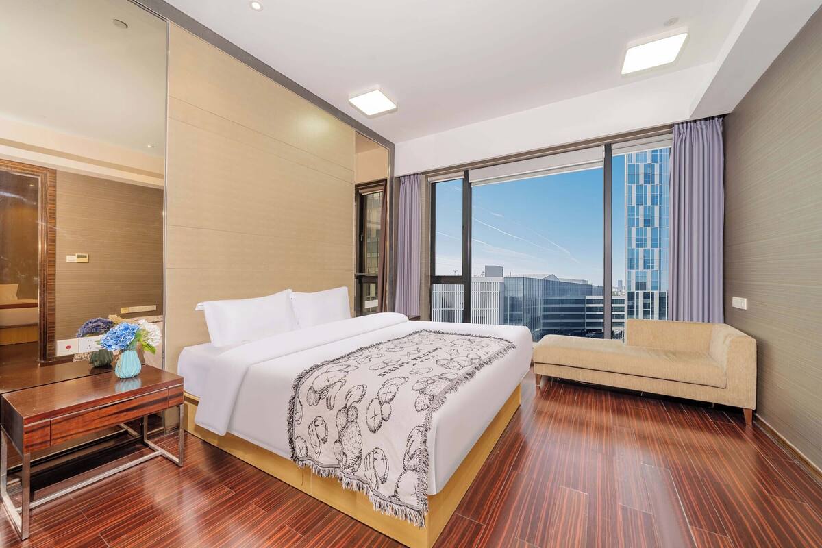 Una habitación de hotel moderna con una cama grande, un sofá, un escritorio y vistas a la ciudad a través de amplios ventanales.