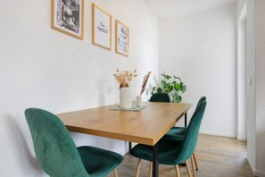 Ein Essbereich mit Holztisch, grün gepolsterten Stühlen, einer Vase mit getrockneten Blumen und gerahmten Bildern an der Wand.