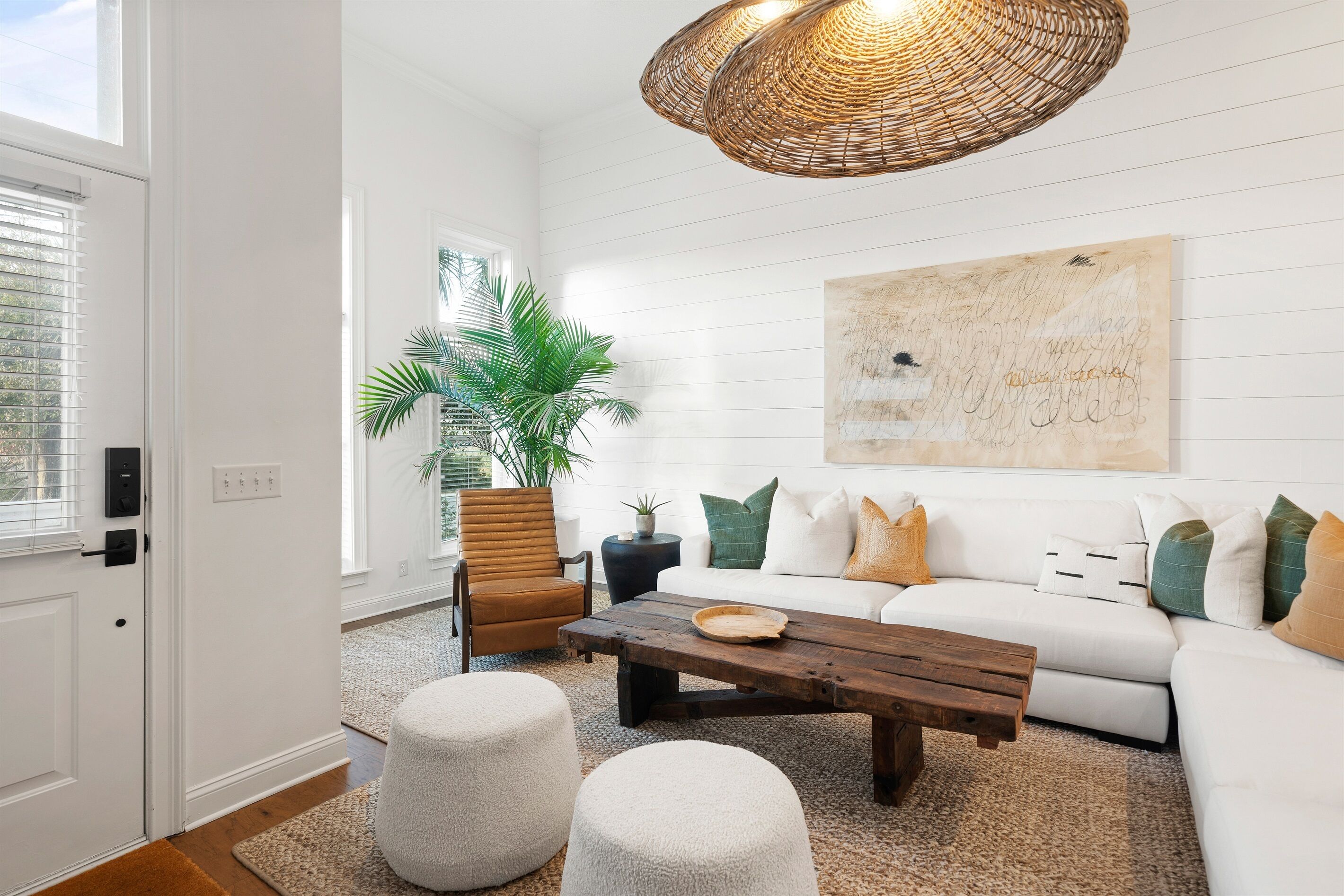 A living room with a white sofa, a wooden coffee table, two white poufs, a wicker pendant light, and a large abstract painting on the wall.