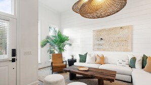 Ein Wohnzimmer mit einem weißen Sofa, einem Holztisch, zwei weißen Poufs, einer Korbtropflampe und einem großen abstrakten Gemälde an der Wand.