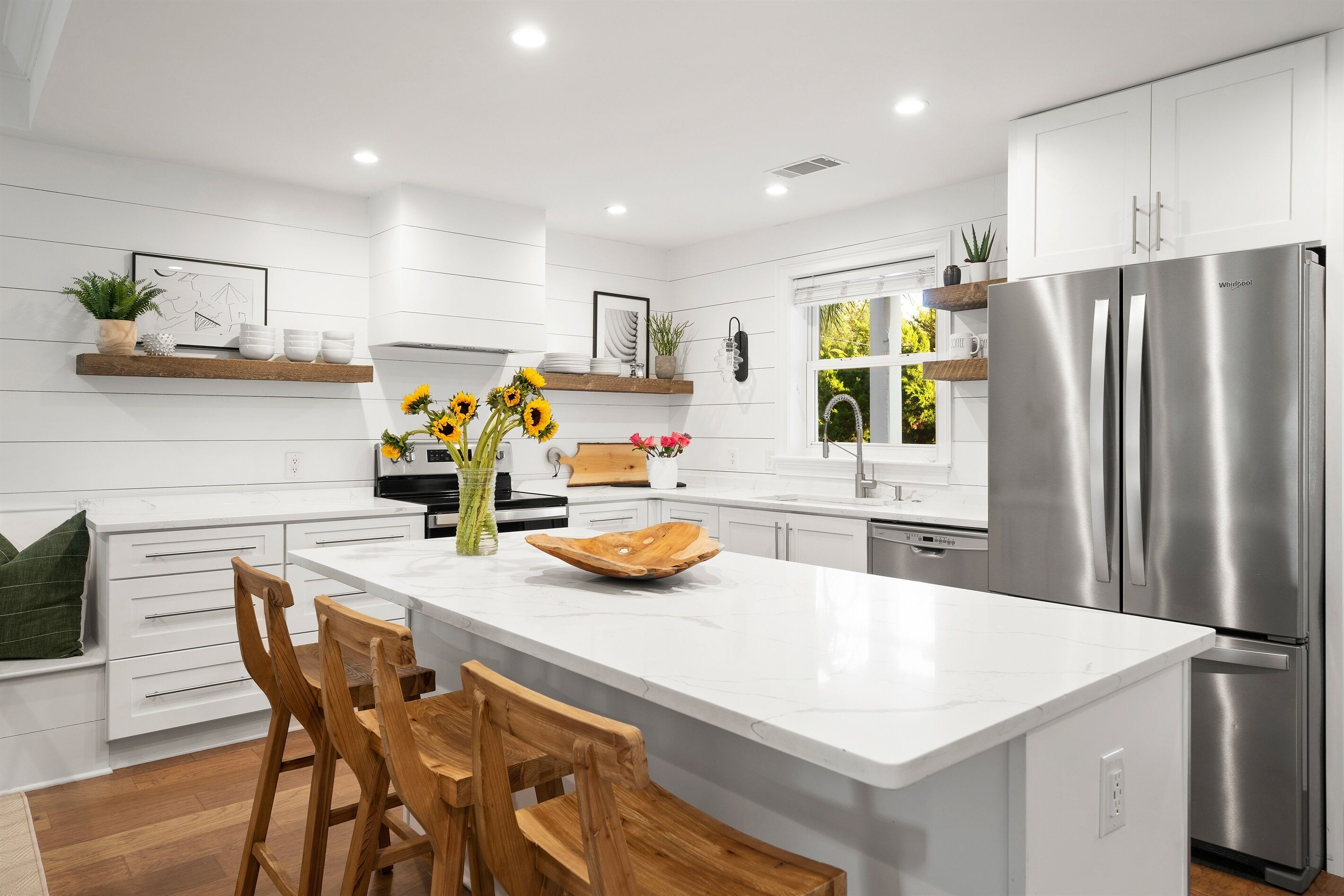 A modern kitchen with white cabinetry, a central island with bar stools, a stainless steel refrigerator, and decorative plants.