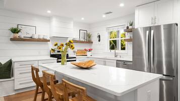 A modern kitchen with white cabinetry, a central island with bar stools, a stainless steel refrigerator, and decorative plants.