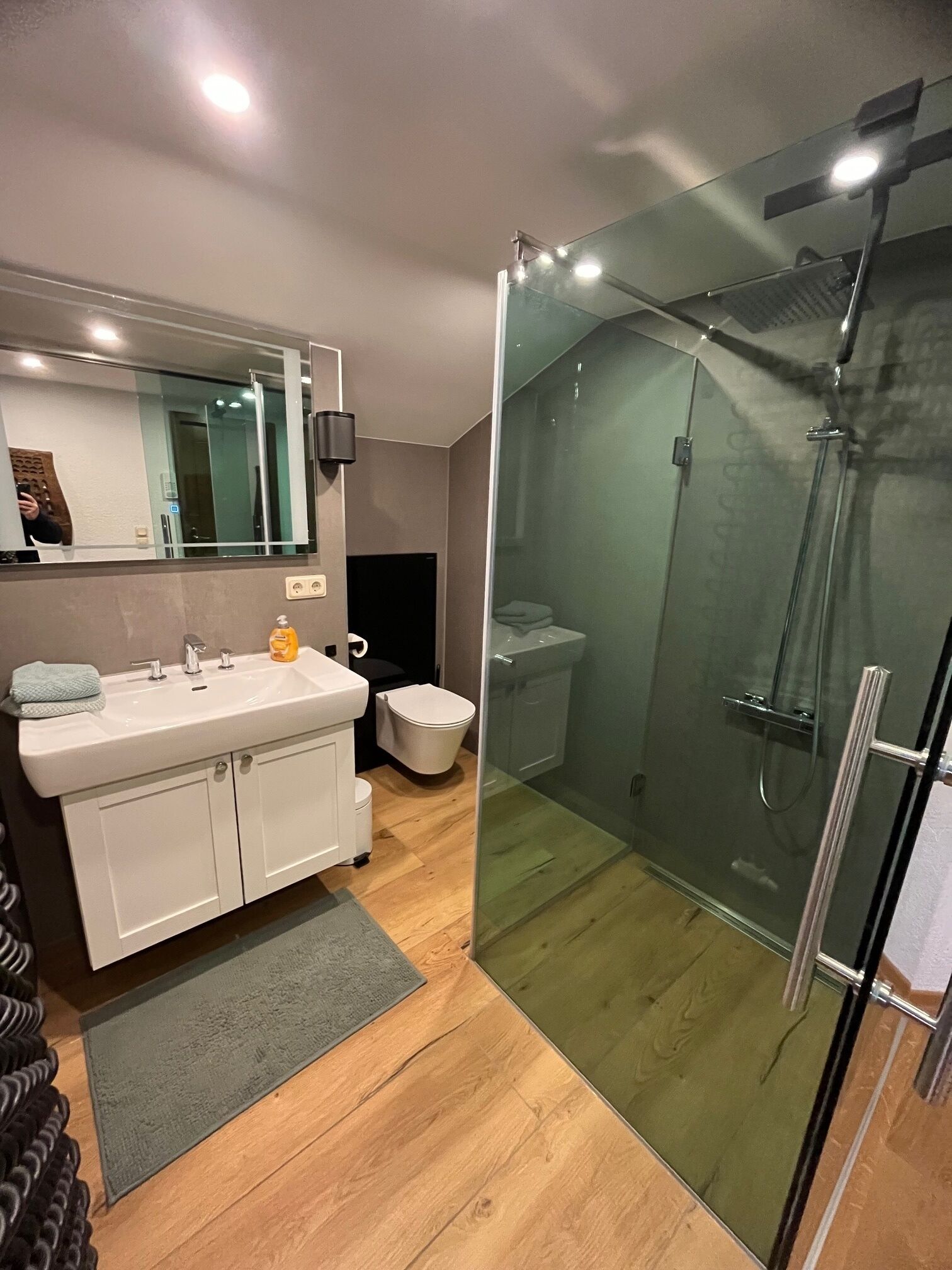 A modern bathroom with a glass-enclosed shower, a white vanity with a mirror, and a wooden floor.
