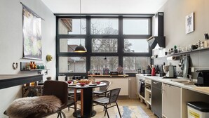 Uma cozinha moderna com janelas amplas, uma área de refeições com mesa redonda e cadeiras, e um lixo visível.