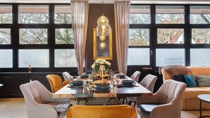 Uma sala de jantar com uma mesa comprida posta para seis pessoas, cercada por cadeiras, um lustre e uma grande janela com cortinas.