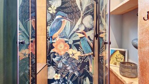 Quarto com porta de vidro, painel de parede com motivos florais e aves, um quadro emoldurado e uma pequena prateleira com uma vela.