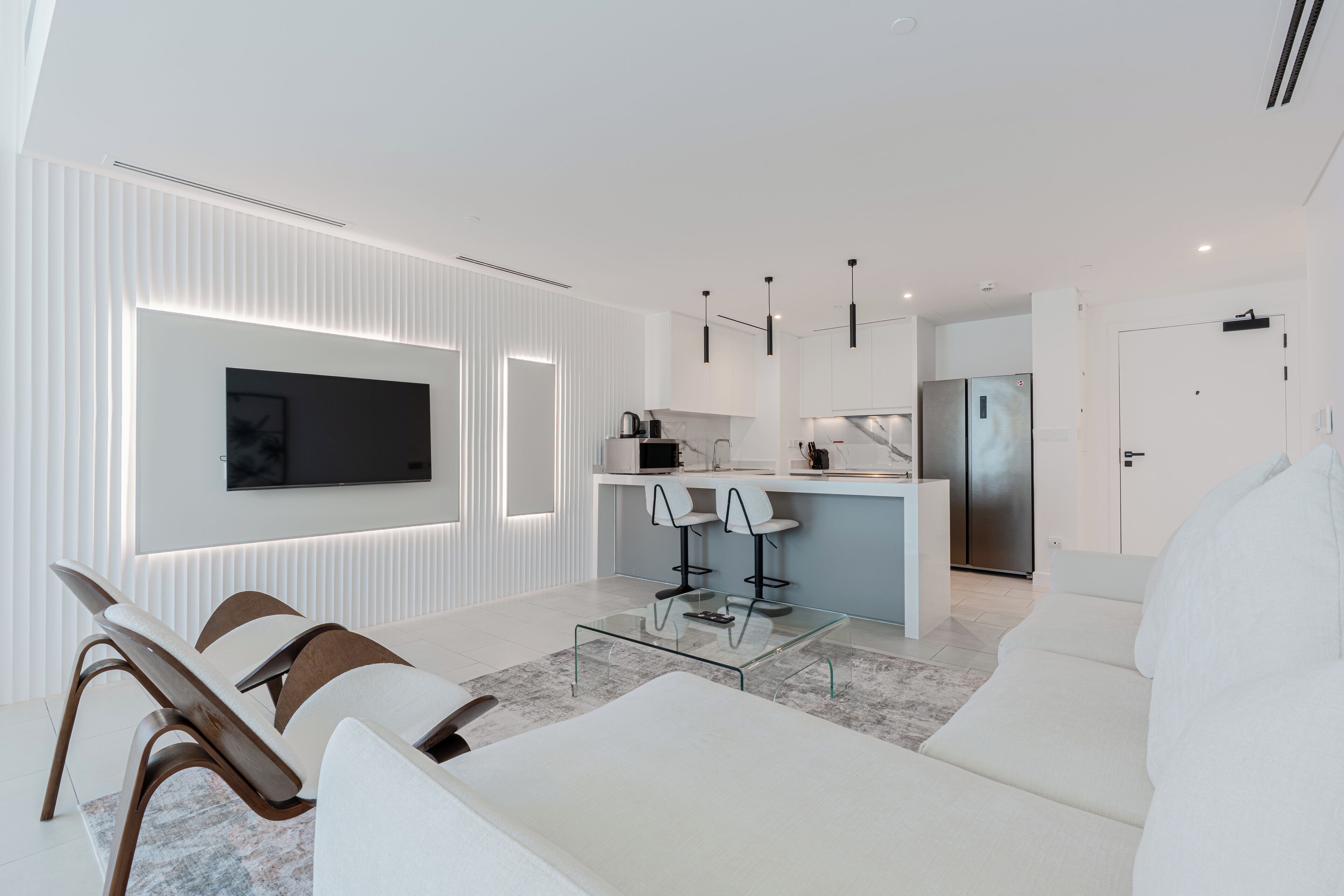A modern living room with a glass coffee table, white sofas, and a TV mounted on the wall.