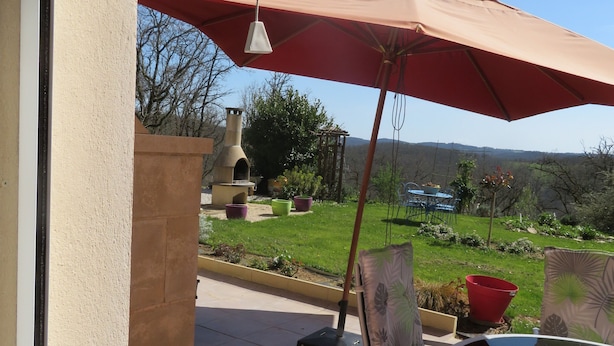 An outdoor patio with a table and chairs, a red umbrella, and a view of the garden and hills. - COLUMBIA (Concorès)