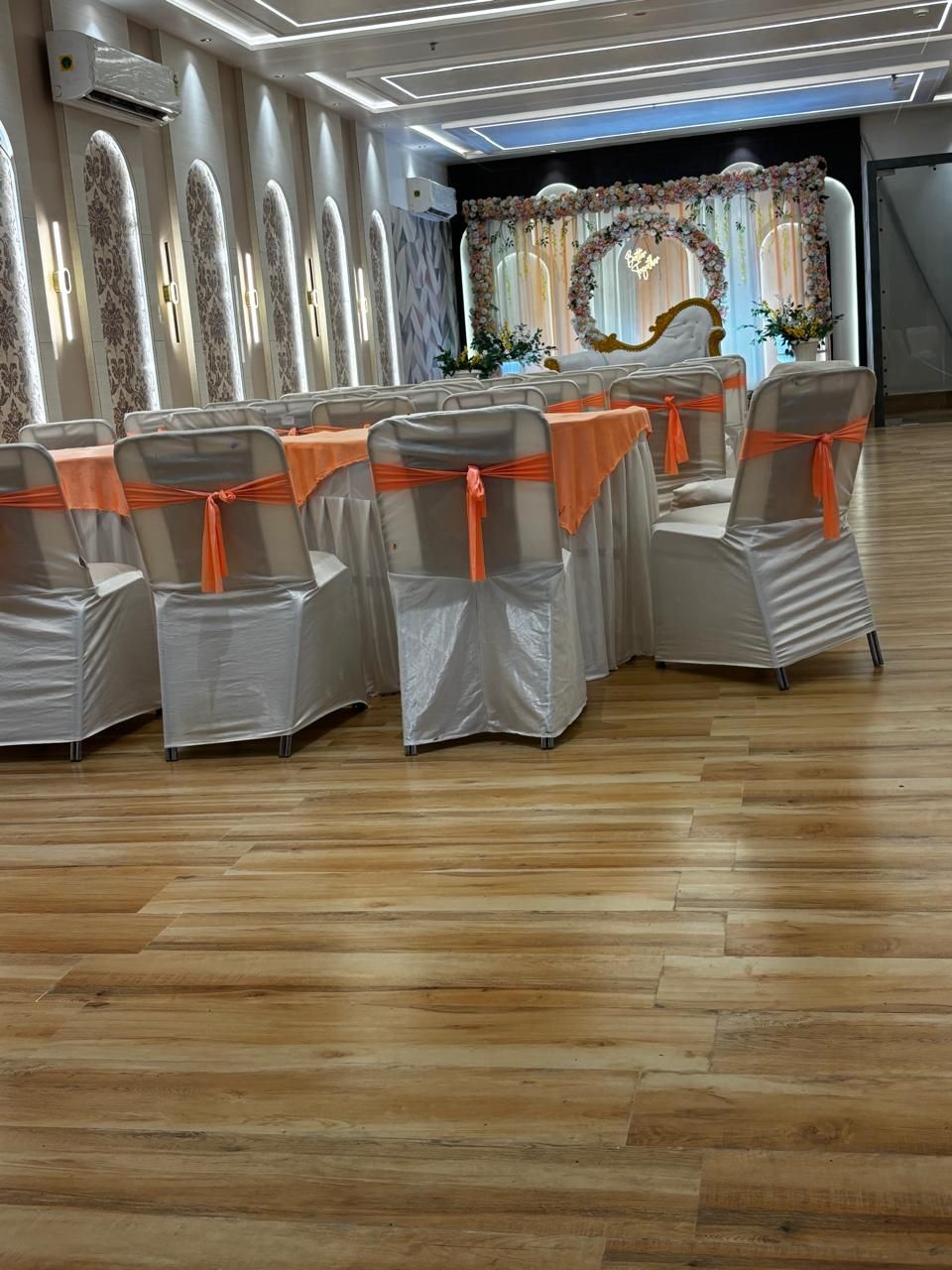 A room with rows of chairs covered in white and orange sashes, wooden flooring, and a floral backdrop.