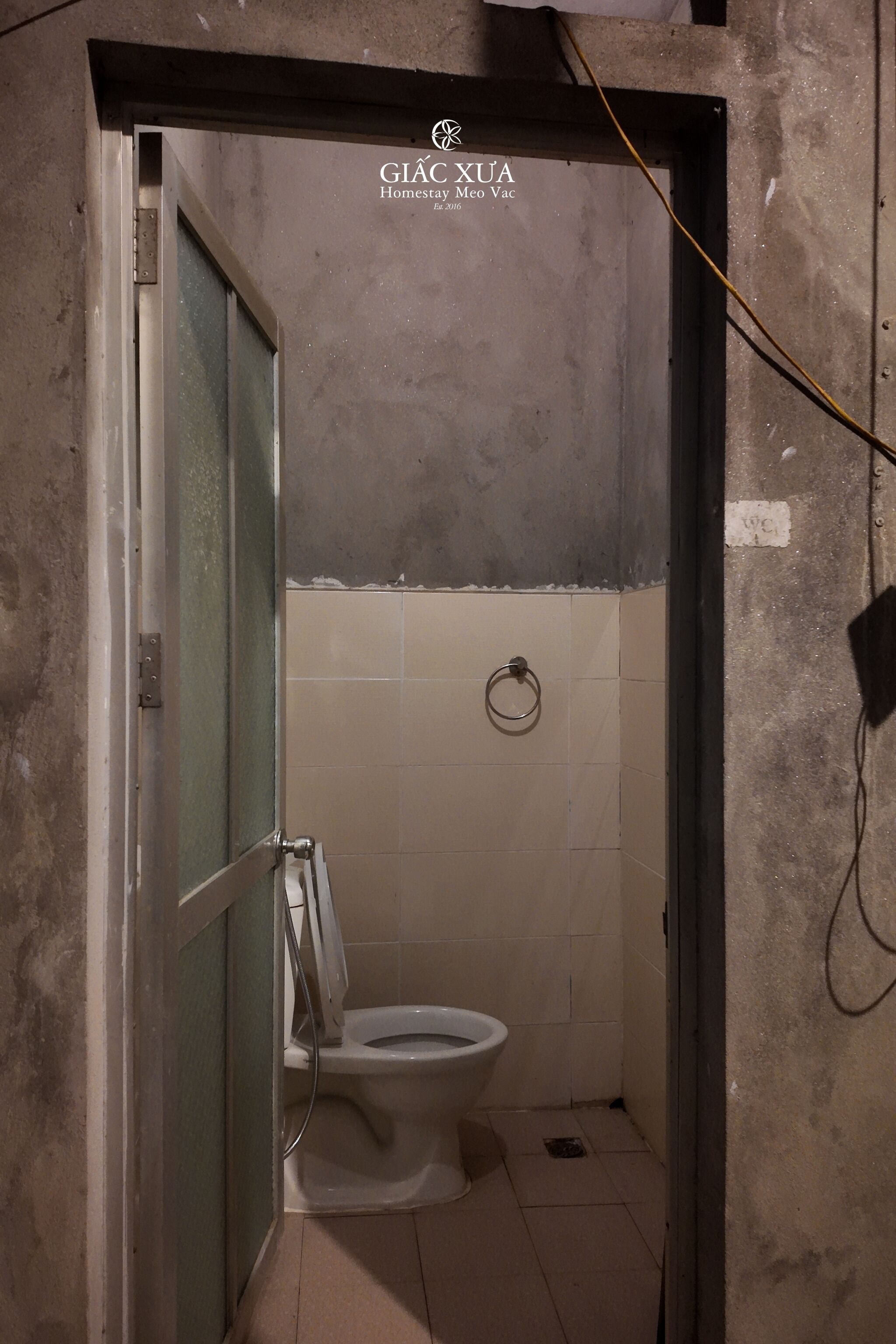 Une petite salle de bain étroite, équipée de toilettes, d’un porte-serviettes et d’un câble électrique apparent.