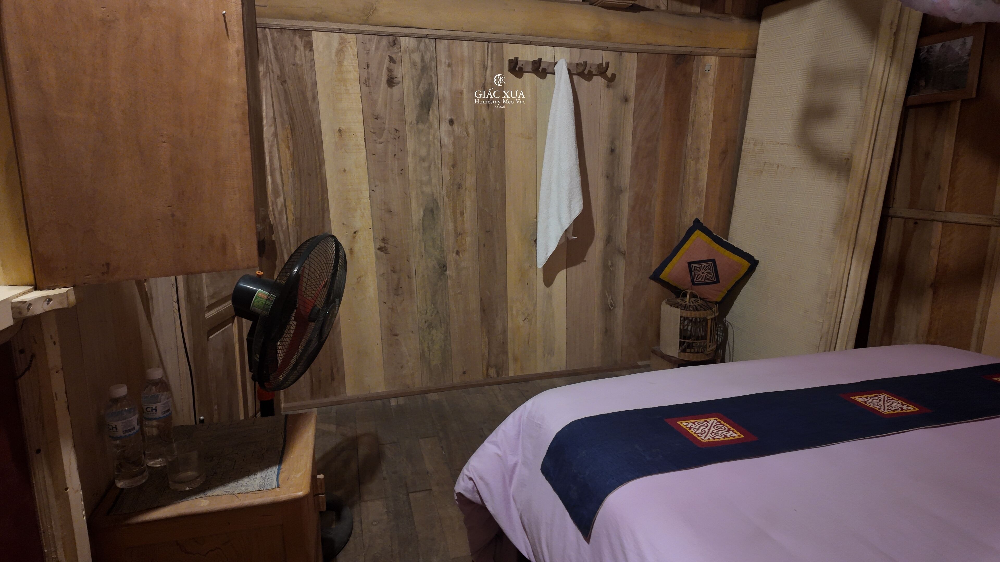 A wooden room with a bed, a fan, a towel, and a small table with bottles.