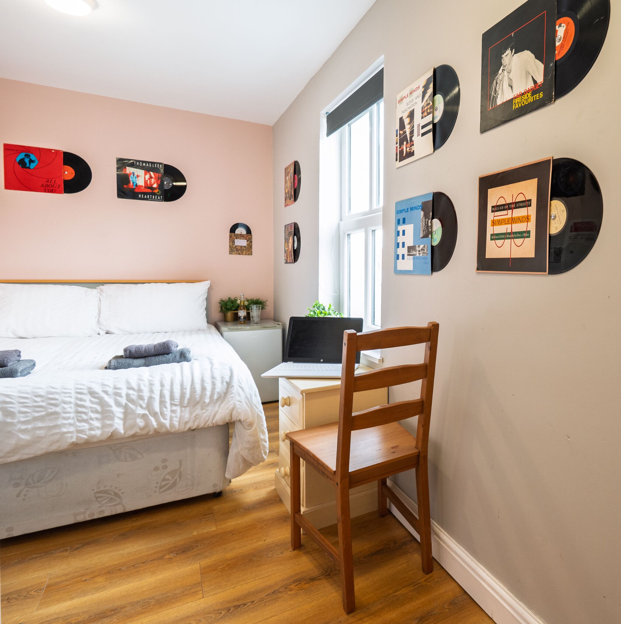 A bedroom with a bed, a wooden chair, a desk with a laptop, and a window.