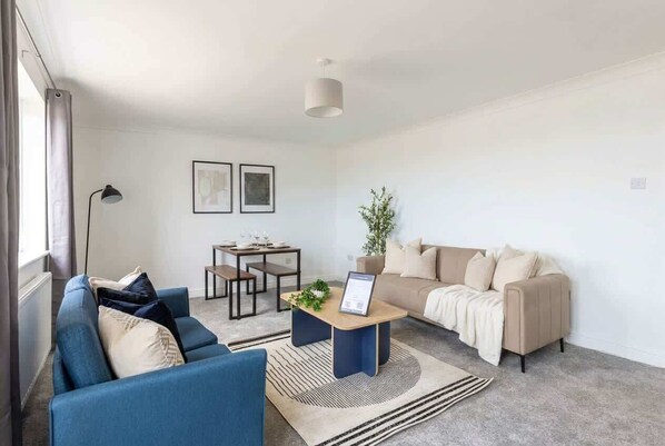 Un salon moderne avec un canapé bleu, un canapé beige et une table basse agrémentée d’une plante. On y trouve également une table à manger avec des chaises et un lampadaire.