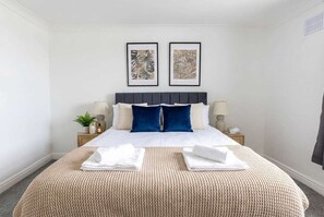 Une chambre d’hôtel bien aménagée, avec un grand lit, deux œuvres d’art encadrées au-dessus de la tête de lit, et une table de chevet avec une lampe et une petite plante.