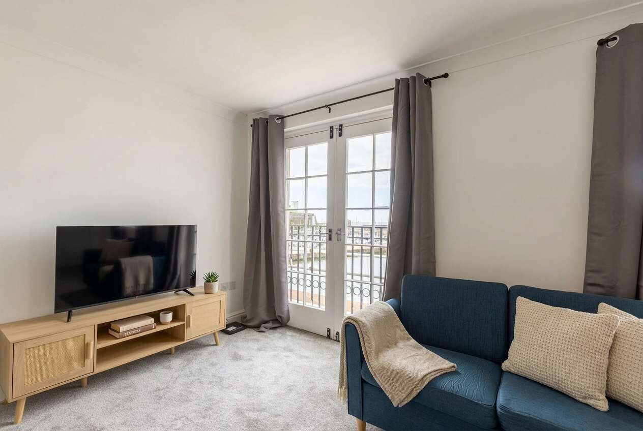 Ein modernes Wohnzimmer mit einem Flachbildfernseher auf einem Holz-TV-Möbel, einem blauen Sofa mit Tagesdecke und einer großen Schiebeglastür mit Vorhängen.
