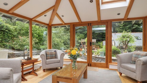 Un salon spacieux avec un plafond haut, des poutres apparentes et de grandes fenêtres donnant sur un jardin.