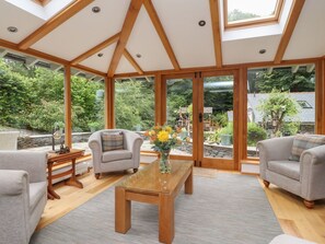 A spacious living room with a high ceiling, wooden beams, and large windows offering a view of a garden. - Bobbin Mill Cottage (Ulverston)