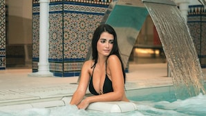 Een vrouw in een zwart zwemkostuum zit bij een zwembad met een fontein.