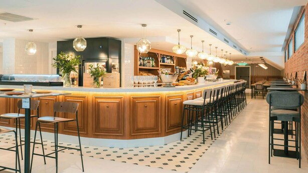 Een moderne bar met een gebogen houten toonbank, meerdere hangende pendelverlichting en een vloer met patroon.
