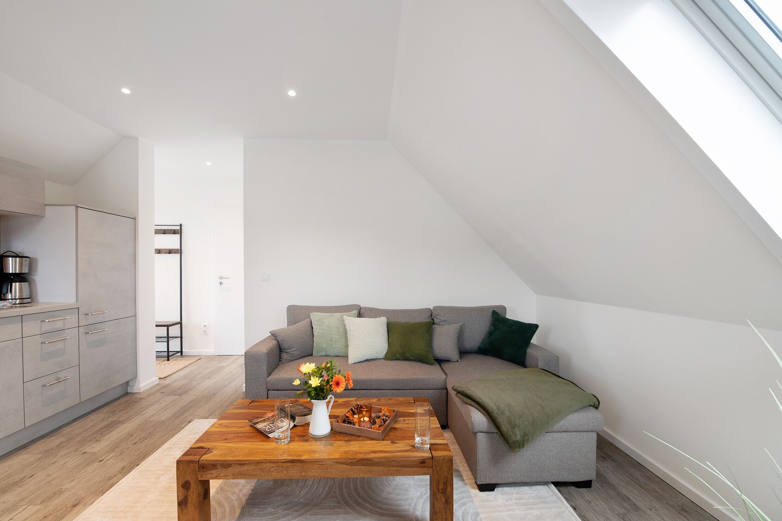A modern loft apartment with a kitchen, a grey sofa with cushions, a wooden coffee table, and a skylight.