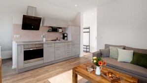 A modern kitchen with a built-in oven and microwave, a grey sofa with cushions, and a wooden coffee table with a vase and flowers.