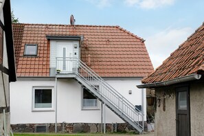 Une maison avec un toit recouvert de tuiles rouges, une façade blanche, une petite fenêtre et un escalier en métal menant à une porte.