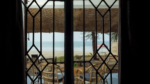 Blick durch ein Fenster mit Metallgitter, der einen Strand mit Stühlen und einem Tisch sowie ein Gebäude mit einem Reetdach zeigt.