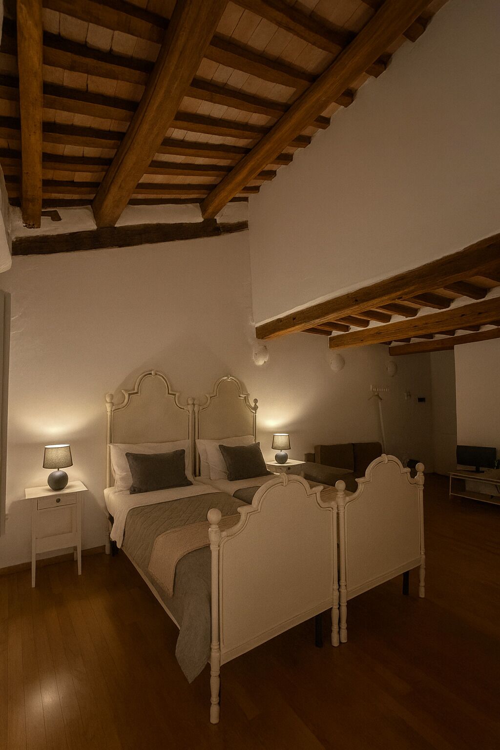 Un dormitori amb sostre de fusta, un llit amb capçalera blanca, dues taules de nit amb làmpades i una televisió sobre un moble.