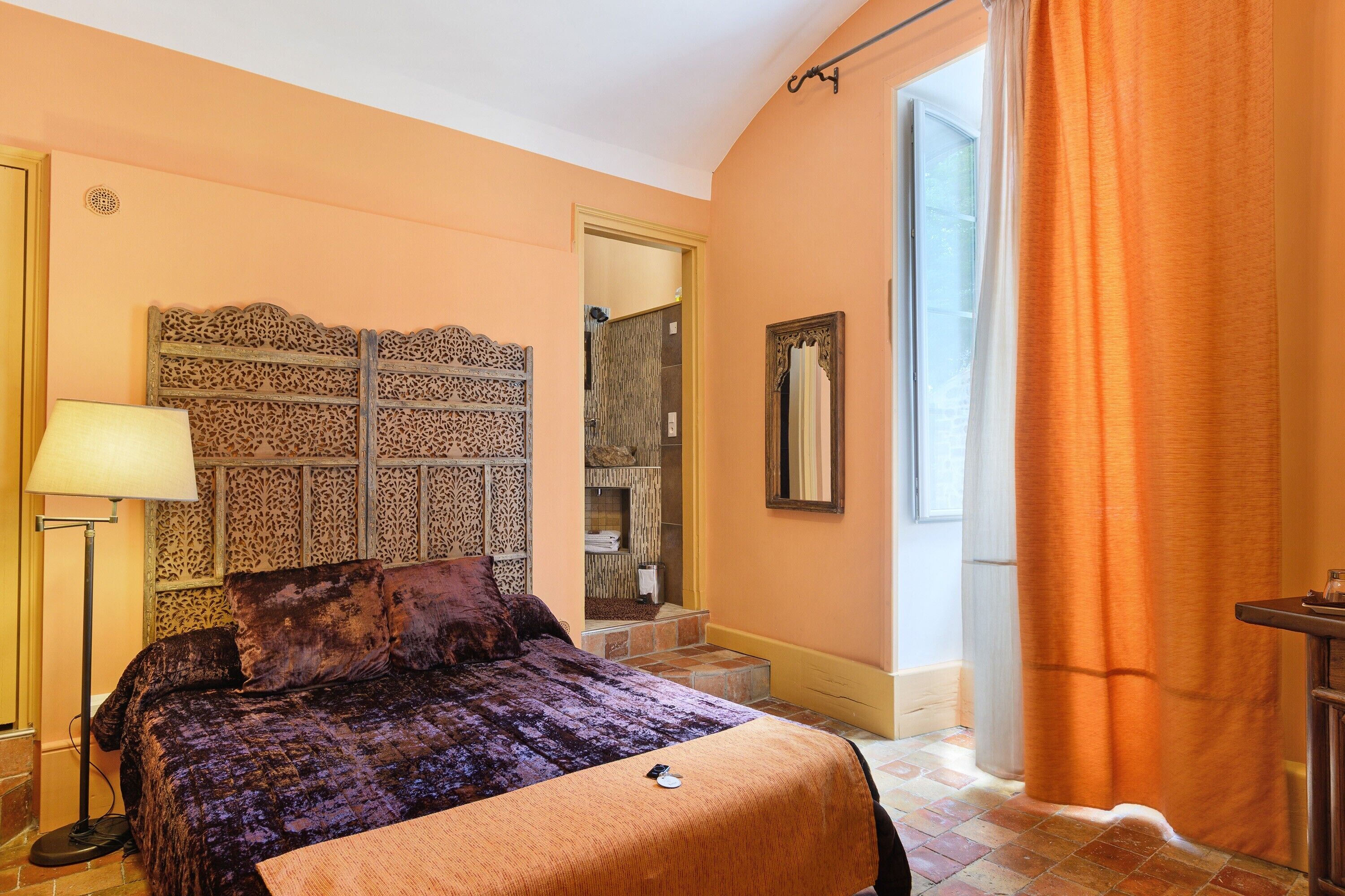 Un dormitorio con cama, lámpara, espejo y ventana con cortinas.