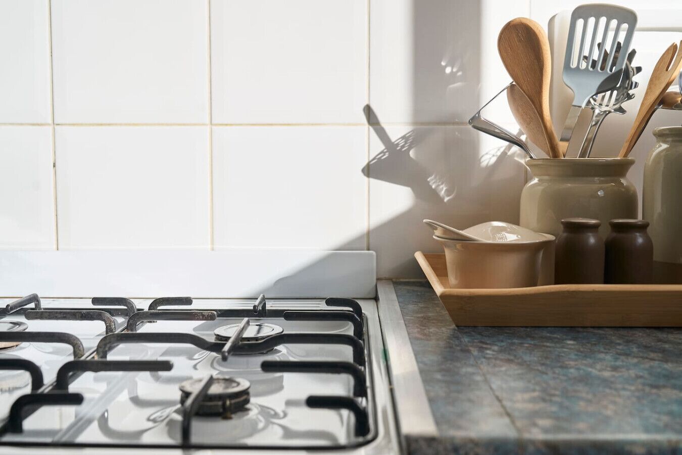 Una cocina con estufa a gas, una bandeja de madera con utensilios de cocina y una ventana iluminada por el sol.