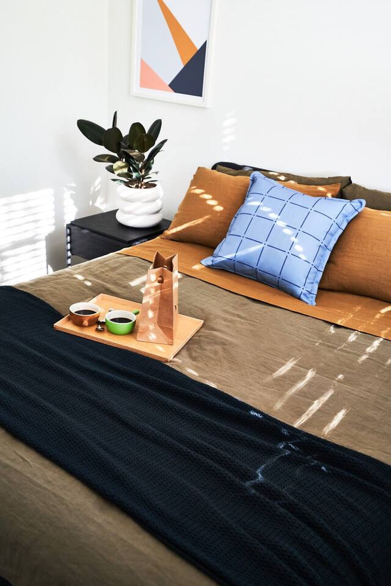 Ein ordentlich gemachtes Bett mit einer braunen Decke, einem blauen Kopfkissen und einem Holztablett mit zwei Tassen Kaffee.