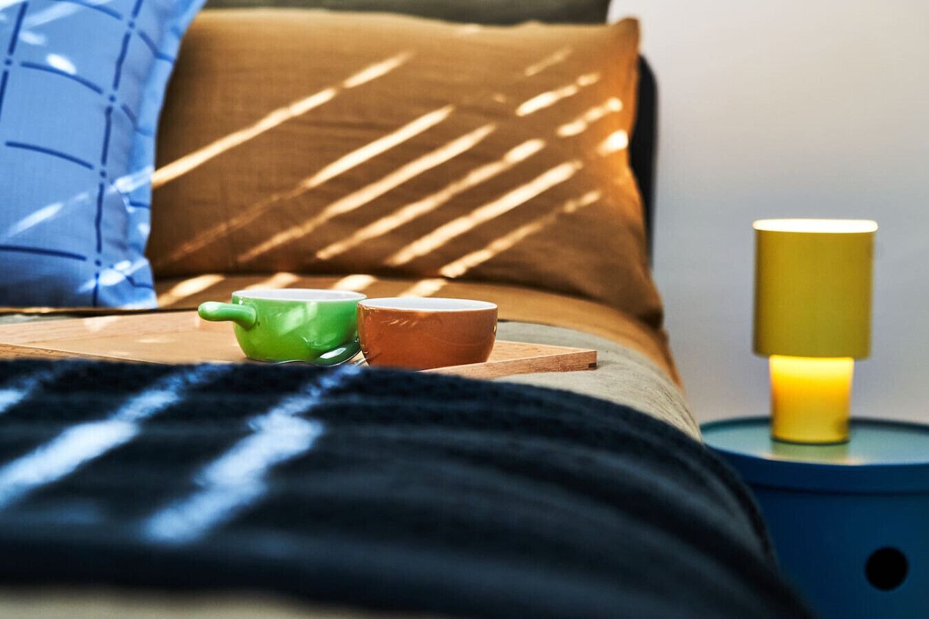 A bed with a wooden headboard, two cups on a wooden tray, and a bedside table with a yellow lamp.