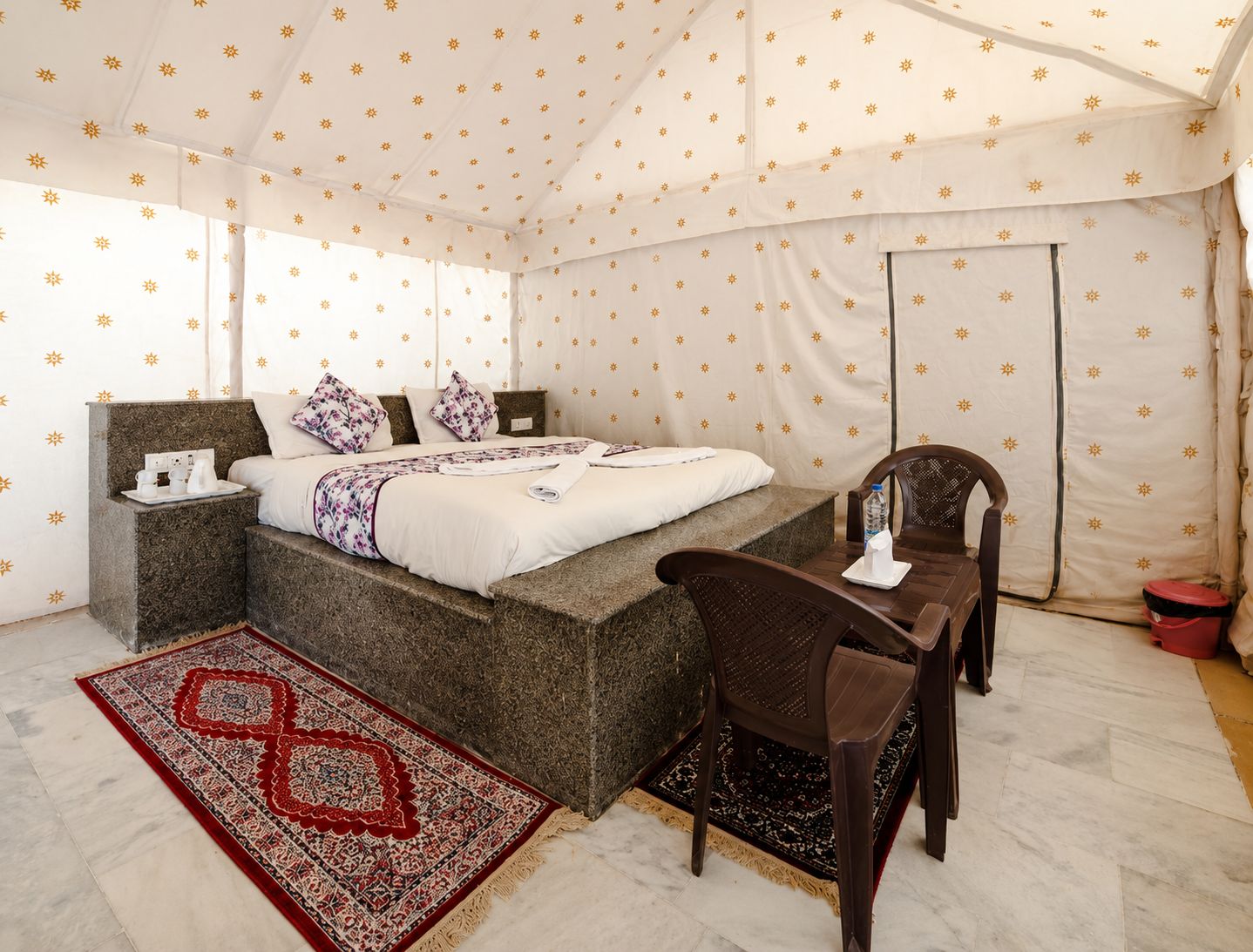 A tented room with a bed, bedside table, chair, and a red patterned rug.