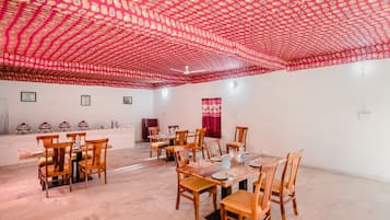A dining area with wooden tables and chairs, a patterned ceiling, and a white wall with a red curtain.