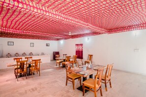 A dining area with wooden tables and chairs, a patterned ceiling, and a white wall with a red curtain. - The Royal Sam Desert Camp (Jaisalmer)