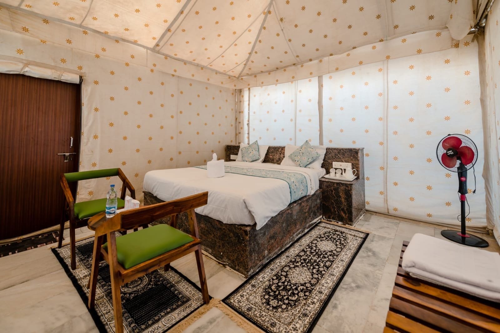 A tented room with a bed, a chair, a bedside table, a fan, and a door.
