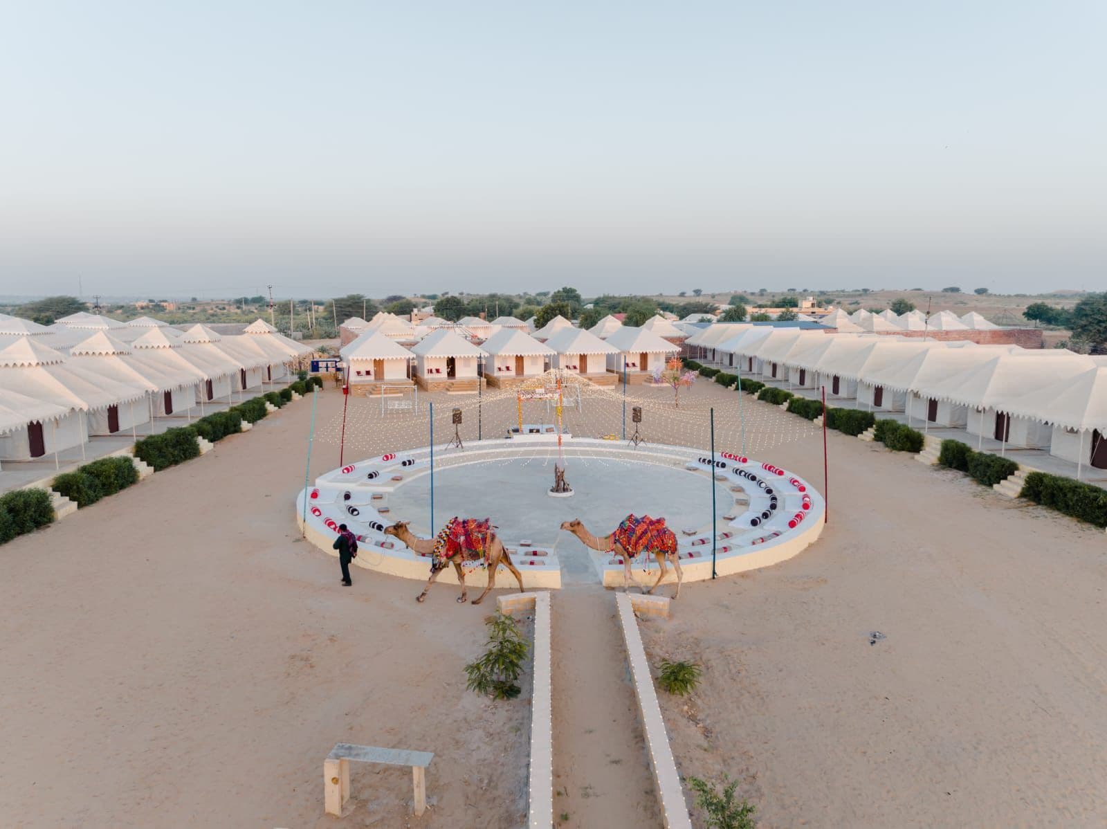 A desert resort with multiple tents arranged in rows, a central circular area with seating, and camels.