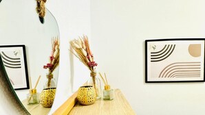 Un espejo refleja una habitación con una mesa de madera, dos cuadros abstractos enmarcados y un jarrón con plantas secas.