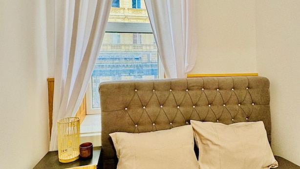 Una habitación con cabecera capitonada, dos almohadas, una mesita de noche con una vela y una ventana con cortinas.