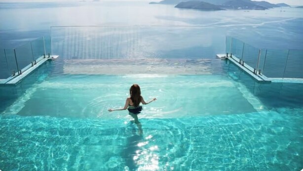 A woman standing in a clear blue infinity pool with a view of the ocean and distant islands.