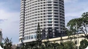 A tall, cylindrical skyscraper with a grid-like facade, surrounded by trees and other buildings.